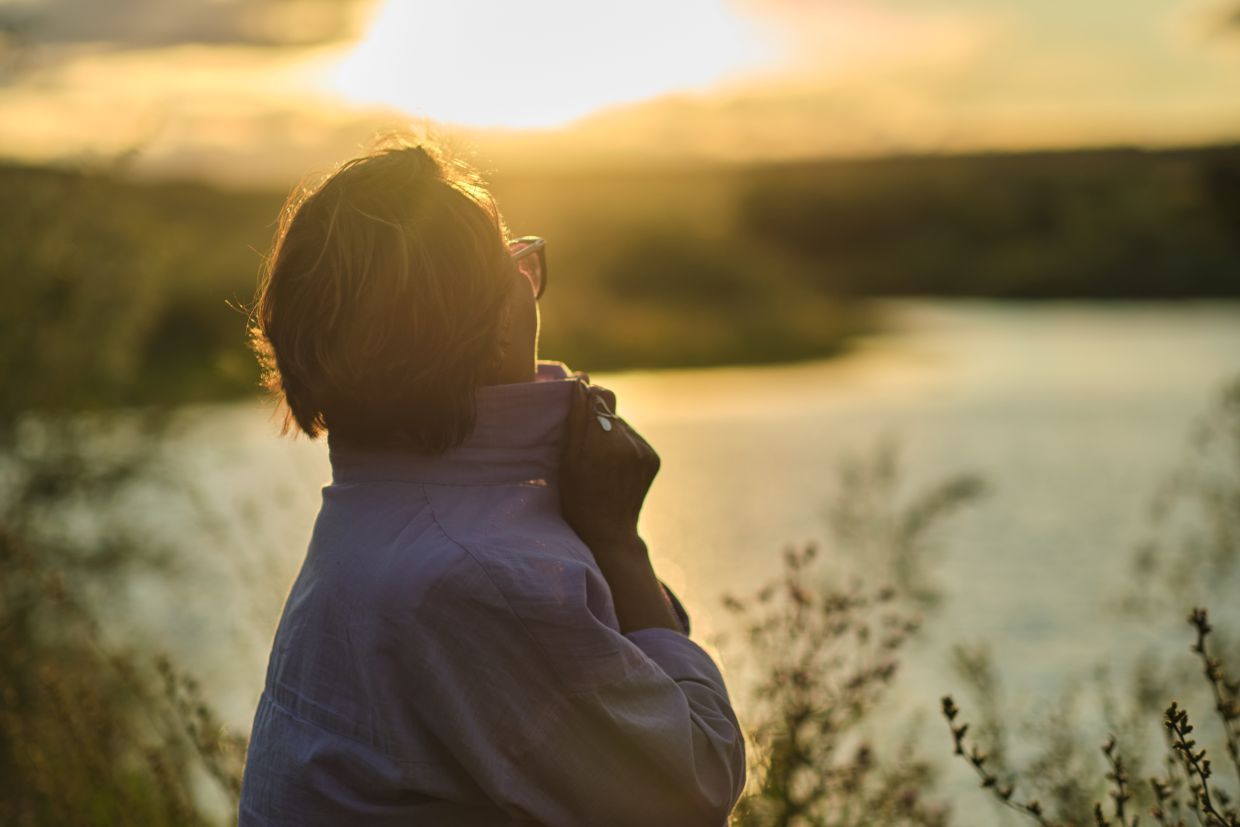 woman standing in a field looking out at sunset over water